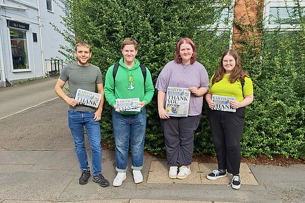 4 Warwick Uni students pictured on a tabloid delivery round in Kenilworth, with a hedge behind them.